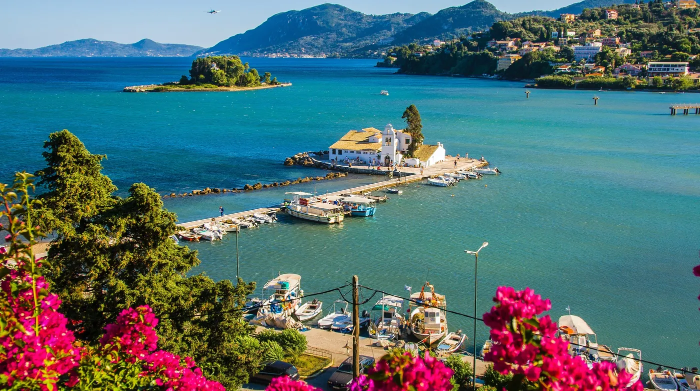A whitewashed monastery at the waterline and a wooded islet, seen through bougainvillea