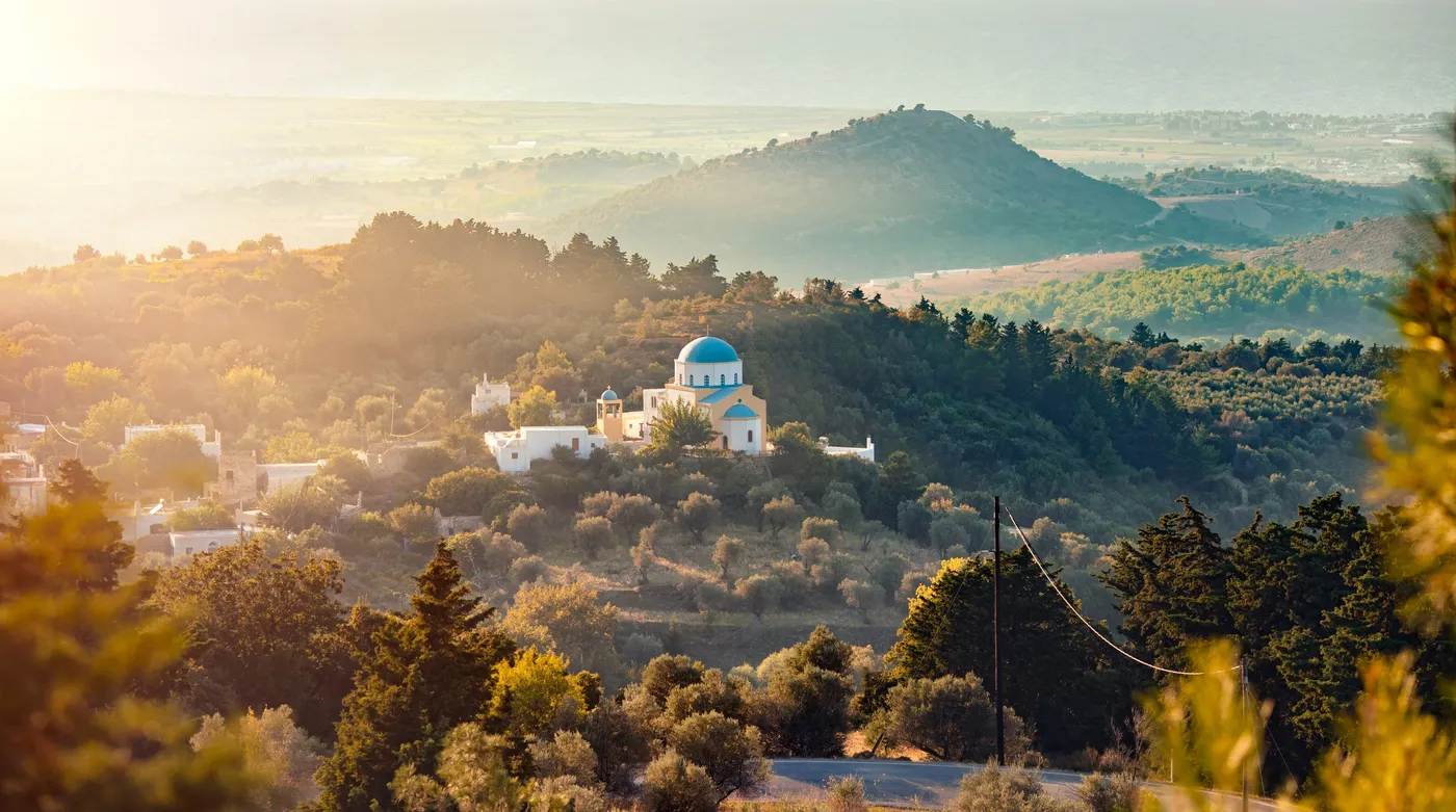 A blue-domed chapel on a hilltop of Kos at sunset, the valley soft below