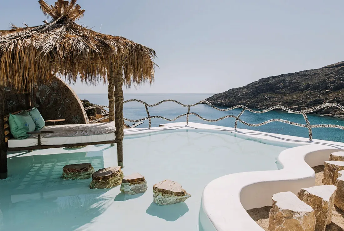 A sculpted white pool with a thatched daybed above a rocky bay