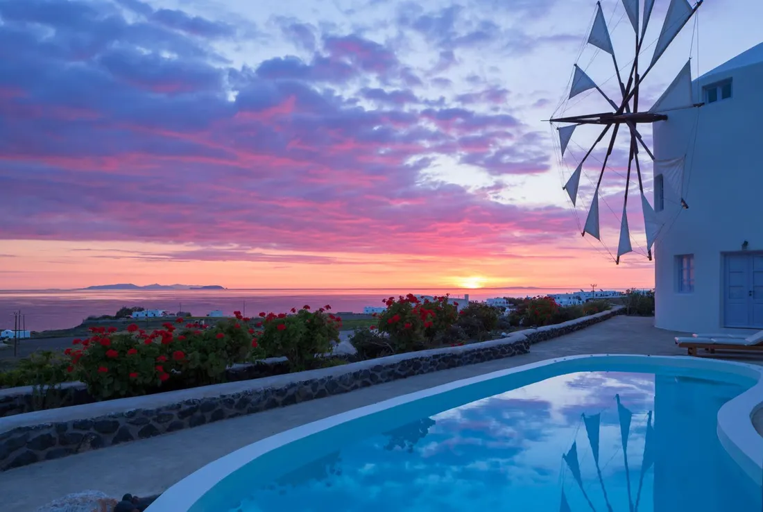 The windmill of Windmill Villas in Santorini at sunset, sails against a pink sky, geraniums and the private pool below