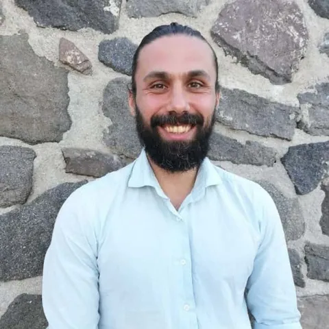Evripidis Mantzaris in a white shirt, smiling, in front of a dry stone wall
