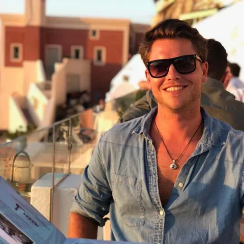Murray Decker in sunglasses and an open collared shirt, smiling, outdoors in front of whitewashed buildings
