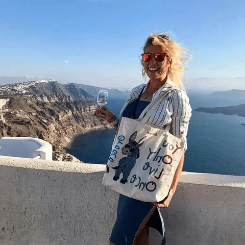 Rose Brown in sunglasses on a clifftop terrace above the sea, a canvas tote over one arm
